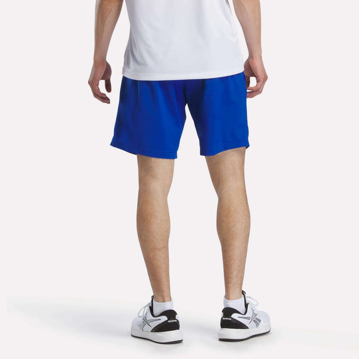 A person stands with their back to the camera, wearing a white shirt, white sneakers with black accents, and blue Reebok Men's Woven Shorts. The background is plain white.