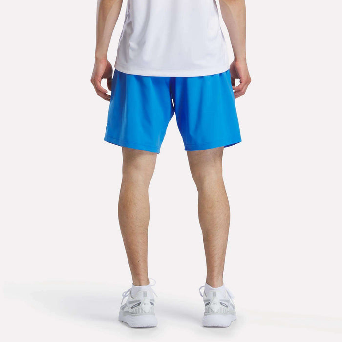 A person stands facing away from the camera, wearing a white shirt, white sneakers, and bright blue Reebok Men's Woven Shorts, against a plain white background.