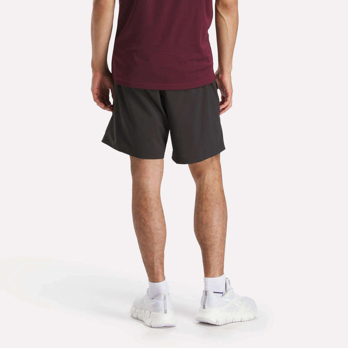A person stands with their back to the camera, wearing a burgundy shirt and Reebok Men's Workout Ready Woven Shorts, known for sweat-wicking comfort, plus white socks and sneakers, against a plain light background.