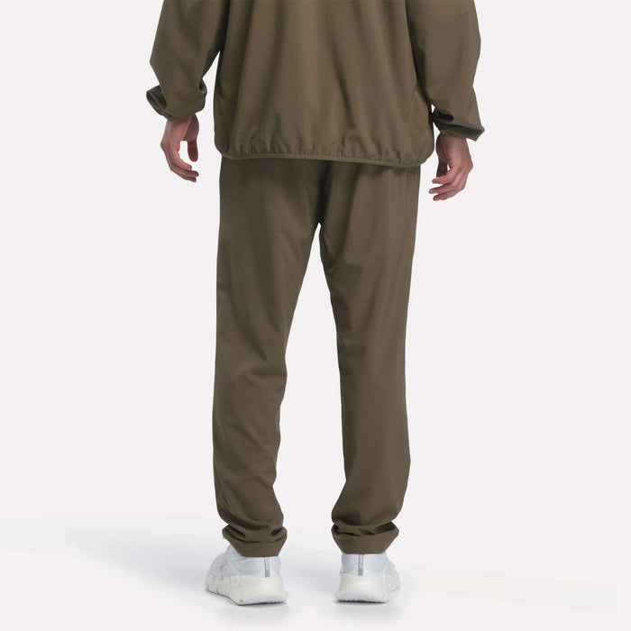 A person stands with their back to the camera, dressed in a loose-fitting olive-green jacket and Reebok Men’s Woven Training Pants, paired with white sneakers, set against a plain white background.