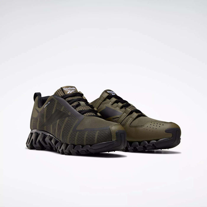 A pair of olive green Reebok Men's ZigWild Trail 6 Shoes with black zigzag soles is displayed on a white background. One shoe stands upright while the other tilts forward, showcasing their trail-inspired design and enhanced cushioning.