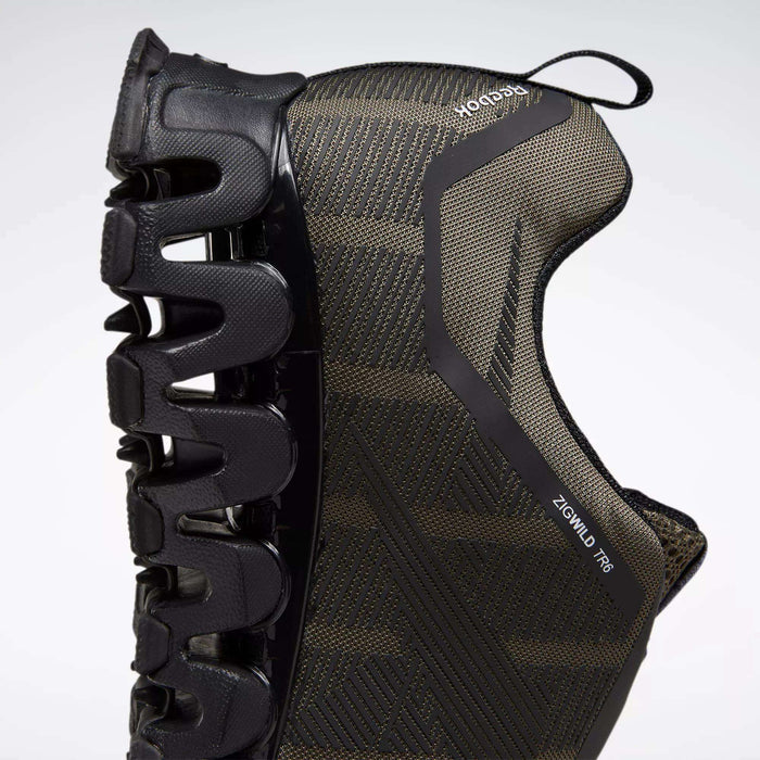 A close-up side view of the Reebok Men's ZigWild Trail 6 Shoes in black, highlighting the unique zigzag-shaped sole for cushioning and a textured upper, set against a light background.