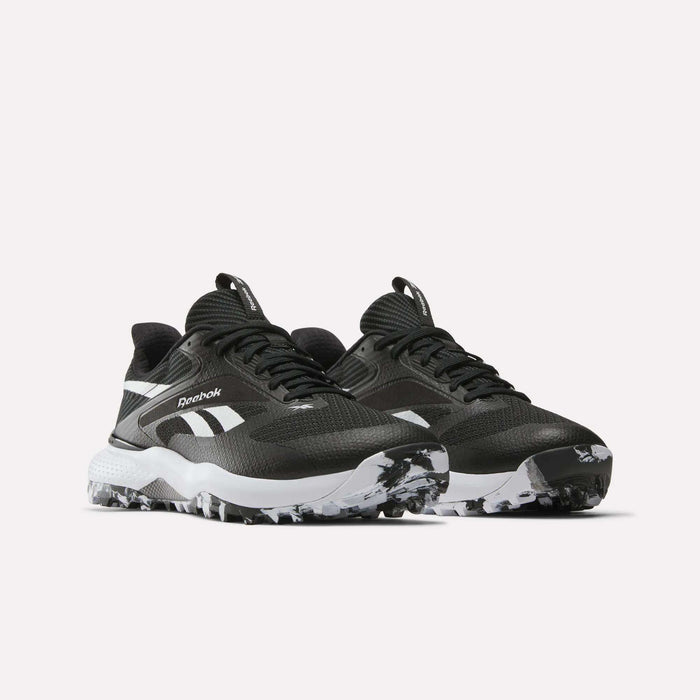 A pair of Reebok Nano Golf Shoes in black with white accents, featuring a FlexWeave upper for durability, a white side logo, and a chunky white sole with black speckles, displayed on a plain light background.