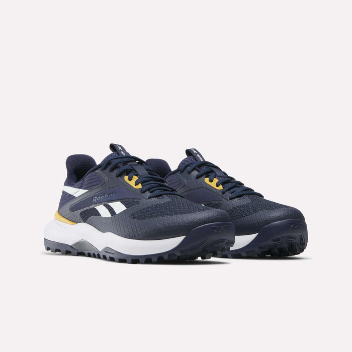 A pair of Reebok Nano Golf Shoes in navy blue with white and yellow accents, featuring a lightweight FlexWeave upper, white soles, and black outsoles, displayed against a plain light background.