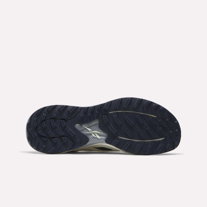 The image displays the dark rubber outsole of the Reebok Nano Golf Shoes, viewed from the bottom. The sole showcases a textured tread pattern and a white Reebok logo at its center, highlighting design elements inspired by lightweight golf shoes that emphasize grip and stability.