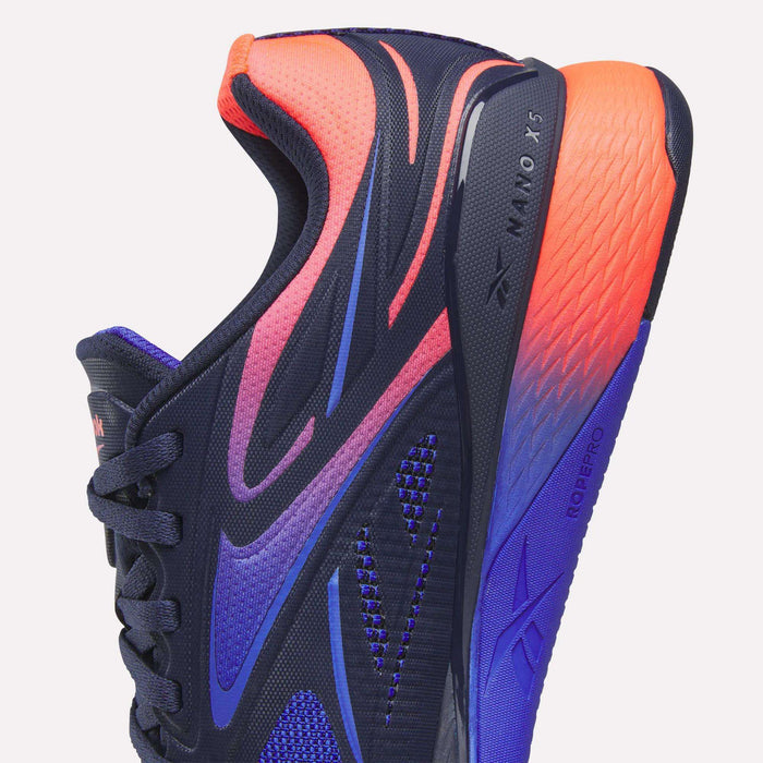 Close-up of Reebok Nano X5 Edge Training Shoes in navy blue with bold purple, pink, and orange gradients. Features textured mesh, black laces, and “Nano X5 Edge” with “RopePro” visible on the side and sole for superior performance.
