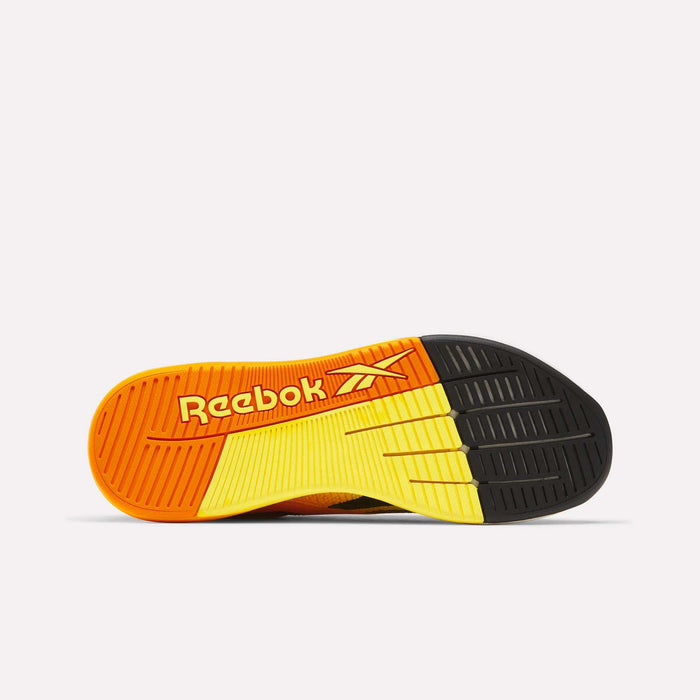 The image displays the sole of Reebok Nano X5 Training Shoes with a gradient design in orange, yellow, and black. The men's shoe has textured grooves for grip and features the orange Reebok logo on the left side.