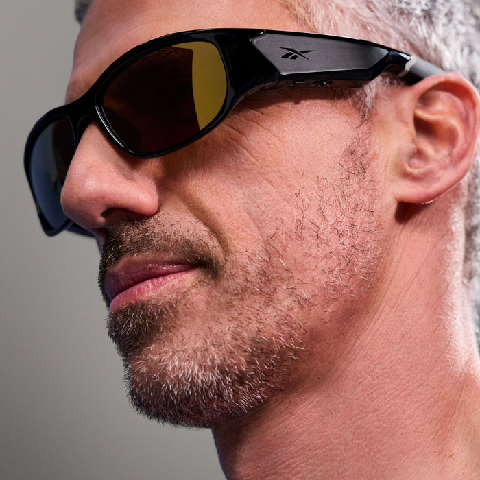 Close-up of a man with short gray hair and stubble, wearing Reebok Nitrous Smart Audio Sunglasses with large tinted lenses. Only the side of his face is visible against a plain gray background.