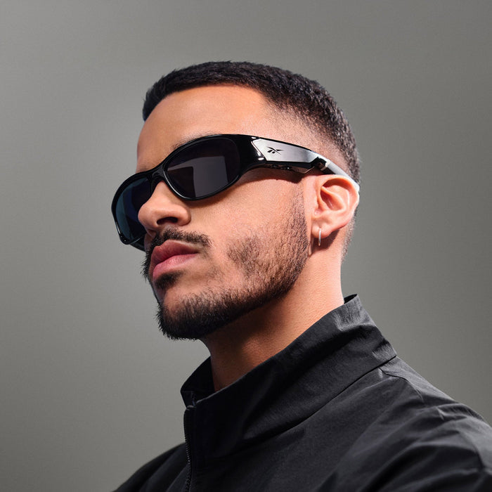 A man with short dark hair, a trimmed beard, and a small hoop earring models the Reebok Nitrous Smart Audio Sunglasses in black, paired with a black jacket, posing against a plain gray background.