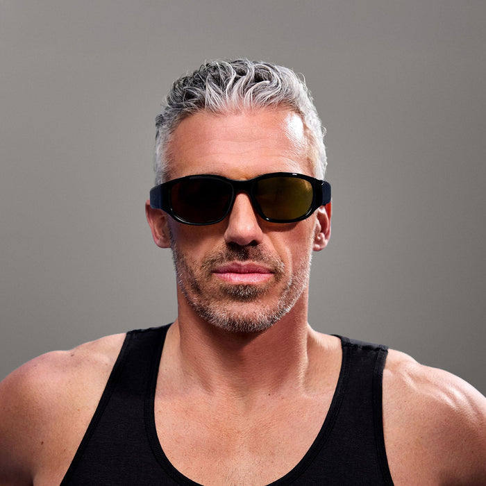 A man with short gray hair and a trimmed beard wears Reebok Nitrous Smart Audio Sunglasses and a black tank top, standing against a plain gray background.