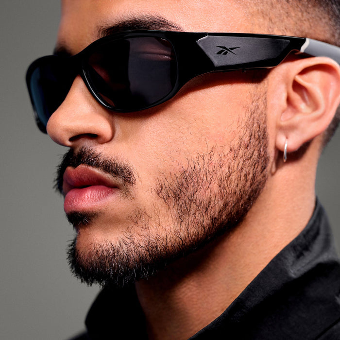 A close-up of a man with a trimmed beard and mustache, wearing Reebok Nitrous Smart Audio Sunglasses with polarized lenses and the Reebok logo visible on the side. He also wears a small hoop earring and a dark collared shirt.