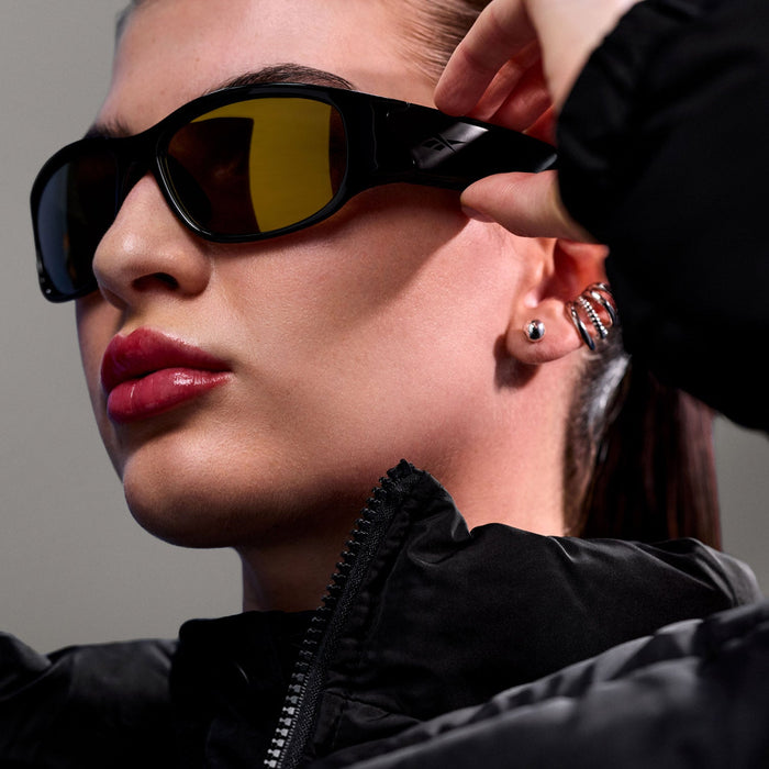 A person in a black zip-up jacket adjusts their Reebok Nitrous Smart Audio Sunglasses. Their hair is tightly pulled back, they wear glossy pink lipstick, and have stud and hoop earrings.