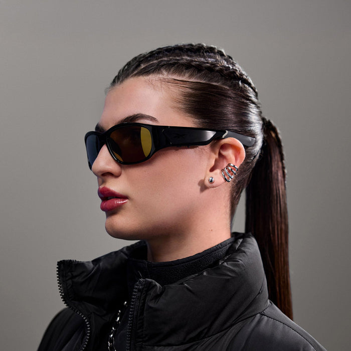 A person with long, slicked-back hair and cornrow braids wears Reebok Nitrous Smart Audio Sunglasses with polarized lenses, a black padded jacket, and multiple earrings, including a colorful ear cuff, against a plain gray background.