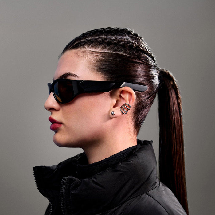 A woman in profile wears Reebok Nitrous Smart Audio Sunglasses with polarized lenses, paired with a black puffer jacket and silver jewelry. Her dark hair is in cornrow braids leading to a ponytail, set against a plain gray background.