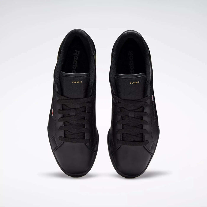 A pair of Reebok Men's NPC II Shoes in black leather is shown from above on a light gray background. These sneakers feature black laces, subtle tongue and side branding, and a classic retro court design.