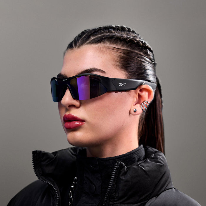 A woman with braided dark hair wears Reebok Octane Smart Audio Sunglasses and a high-collared black jacket, paired with bold earrings and glossy lipstick, standing against a plain gray background.