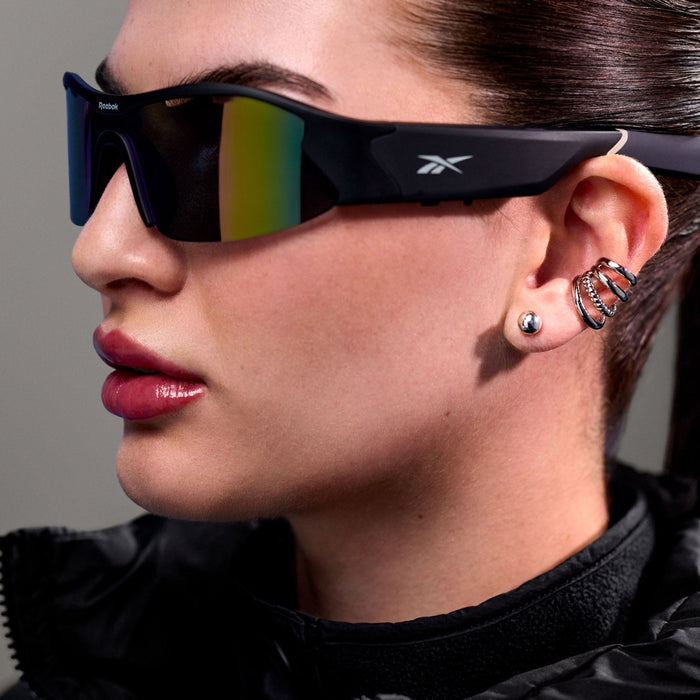 A person wearing large Reebok Octane Smart Audio Sunglasses, a black jacket, and several silver earrings—including a spiral-shaped one—is shown in close-up from a side angle.