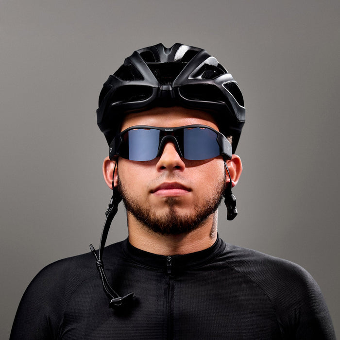 A man wearing Reebok Octane Smart Audio Sunglasses with polarized lenses, a black cycling helmet, and a black jersey stands against a gray background, looking straight ahead with a neutral expression.