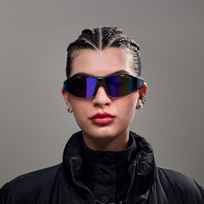 A person with cornrow braids wears Reebok Octane Smart Audio Sunglasses with polarized lenses, a black high-collared jacket, and earrings while posing against a plain gray background.