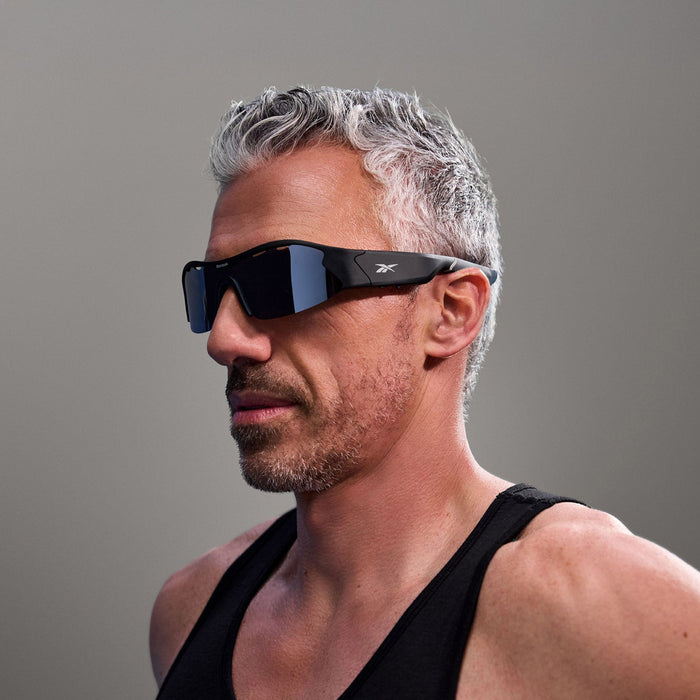 A man with short gray hair and a trimmed beard wears large, black Reebok Octane Smart Audio Sunglasses with polarized lenses and a black tank top against a plain, neutral background.
