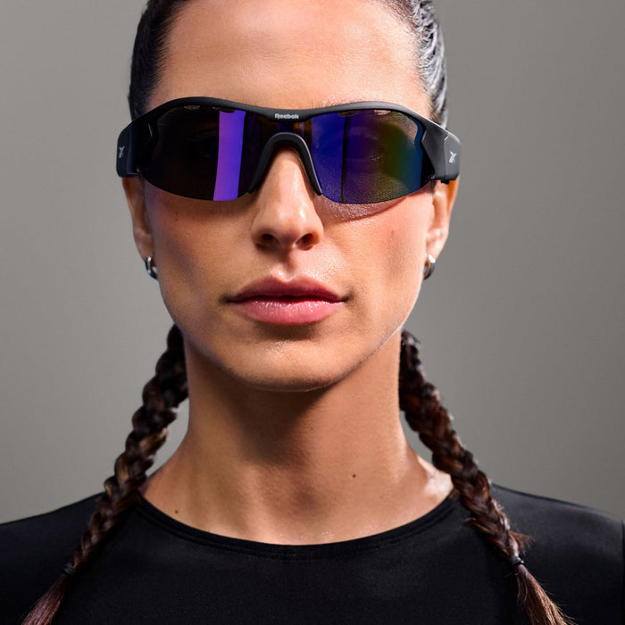 A person with braided hair wears Reebok Octane Smart Audio Sunglasses and a black top, looking directly at the camera against a plain gray background.