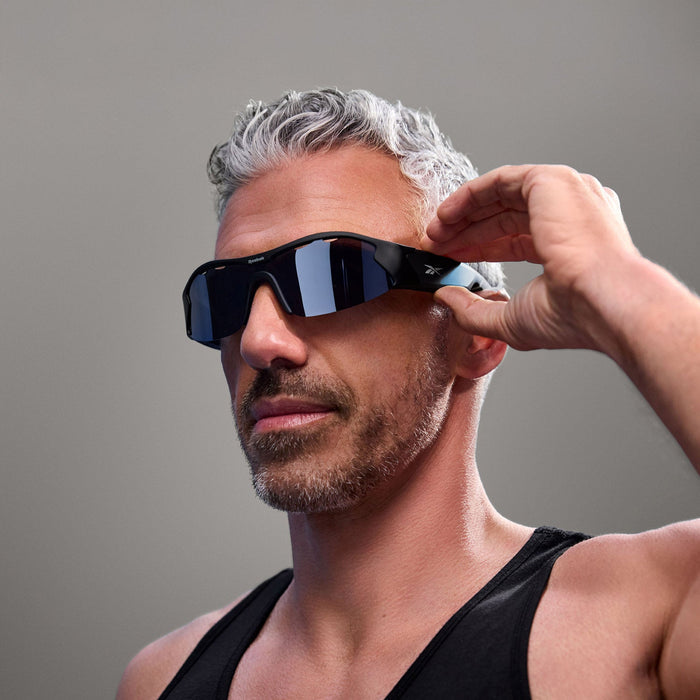 A man with short gray hair and a beard wears Reebok Octane Smart Audio Sunglasses with polarized lenses, dressed in a black tank top and touching the side of the glasses against a plain gray background.