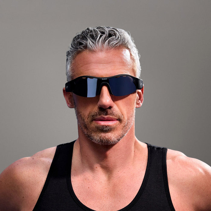 A middle-aged man with short gray hair and a trimmed beard wears Reebok Octane Smart Audio Sunglasses and a black tank top, standing against a plain gray background.