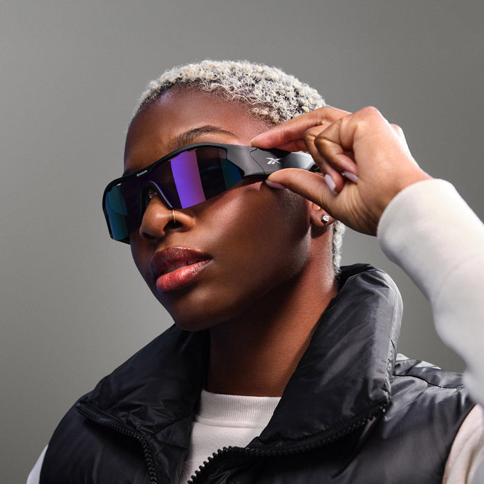 A person with short, curly blond hair adjusts Reebok Octane Smart Audio Sunglasses—large, futuristic Bluetooth sport eyewear—while wearing a black puffer vest over a white top against a neutral gray background.
