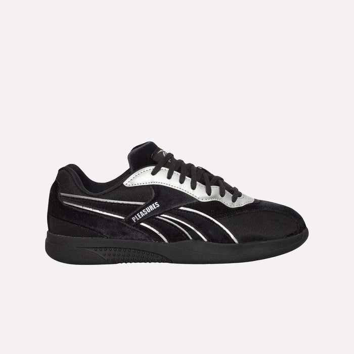The Reebok Pleasures Hammer Street Shoes, a black sneaker with a velvet upper and white and silver accents, features "PLEASURES" on the side—part of the PLEASURES collaboration—set against a plain light blue background.