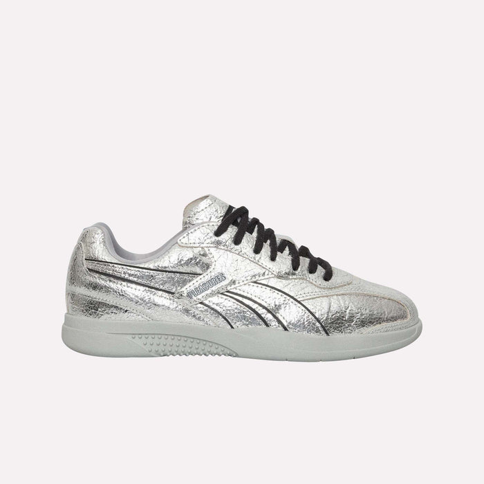 The Reebok Pleasures Hammer Street Shoes feature a silver metallic finish with black laces, a textured surface, and a light-colored sole—offering bold street style in a modern silhouette, as presented on a simple light backdrop.