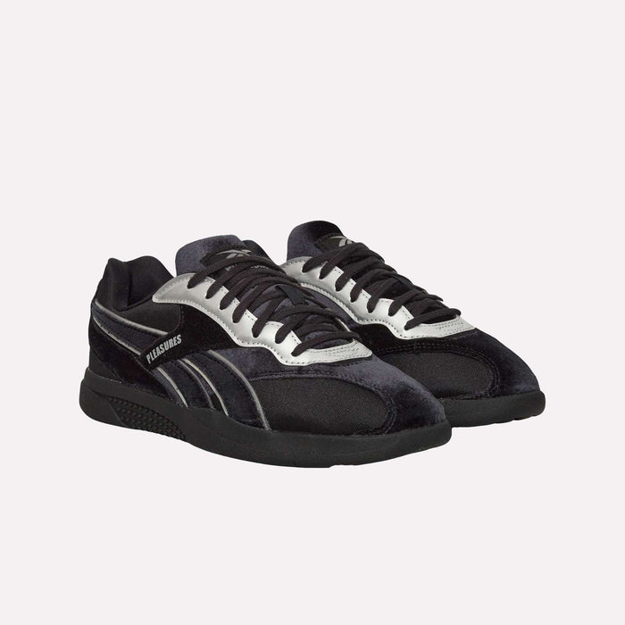 A pair of Pleasures Hammer Street Shoes by Reebok in black and silver, featuring PLEASURES branding on the side and a sleek velvet upper, displayed against a plain white background.