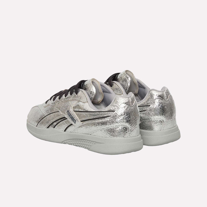 A pair of Pleasures Hammer Street Shoes by Reebok, featuring silver metallic tones with black accents, are displayed from the back and side on a plain light background—the distinctive design evokes a subtle grunge sneakers vibe.