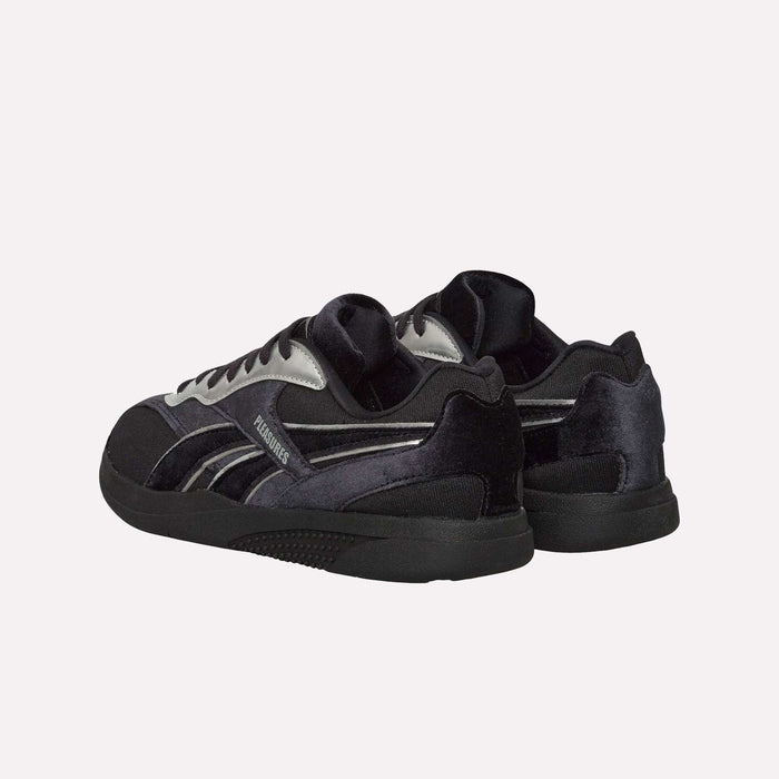 A pair of Pleasures Hammer Street Shoes by Reebok, featuring a black and gray color scheme inspired by classic Reebok indoor soccer shoes, is shown on a white background, positioned at an angle with the heels slightly facing forward.