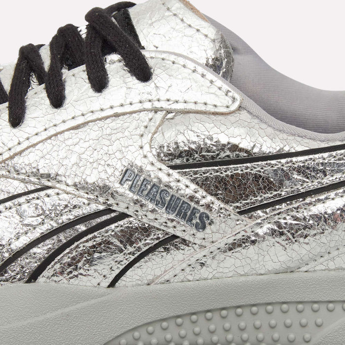 Close-up of the Pleasures Hammer Street Shoes by Reebok, featuring a silver cracked-texture upper, black laces, and PLEASURES branding on the side. Inspired by grunge streetwear, these sneakers incorporate bold black stripe details and a textured gray sole, showcasing a striking collaboration reminiscent of classic Reebok indoor soccer shoes.
