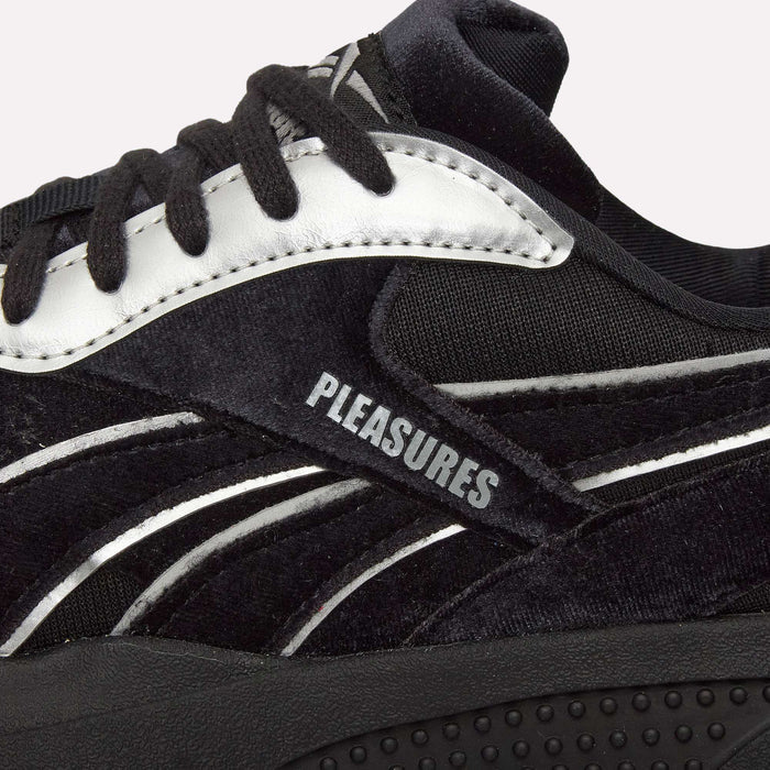 Close-up of the Pleasures Hammer Street Shoes by Reebok, featuring a black velvet upper with white accents, black laces, and “PLEASURES” printed in bold white uppercase letters on the side. This collaborative design draws inspiration from classic Reebok indoor soccer shoes.