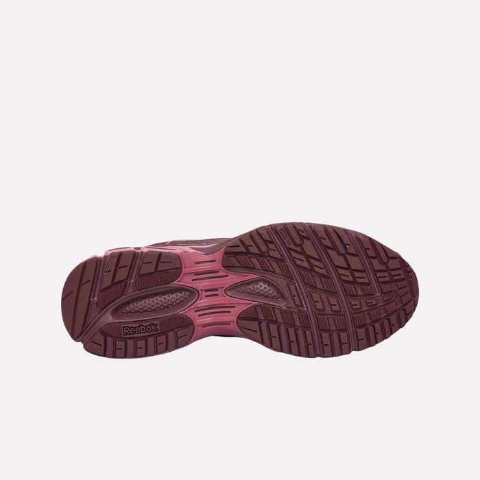 The image displays the maroon rubber outsole of the Reebok Premier Road Ultra Shoes, highlighting textured tread patterns for optimal grip and a visible logo near the heel. Drawing inspiration from an archival running silhouette, this model from Reebok is engineered for enhanced comfort with its dual-density midsole. The background features a plain light pink color.
