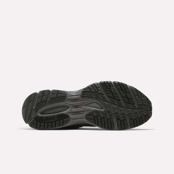 The image shows the black rubber outsole of Reebok Premier Trinity Shoes, viewed from the bottom. The tread has patterns and grooves for traction and flexibility, resembling retro running shoes. The "Reebok" logo appears on the sole.
