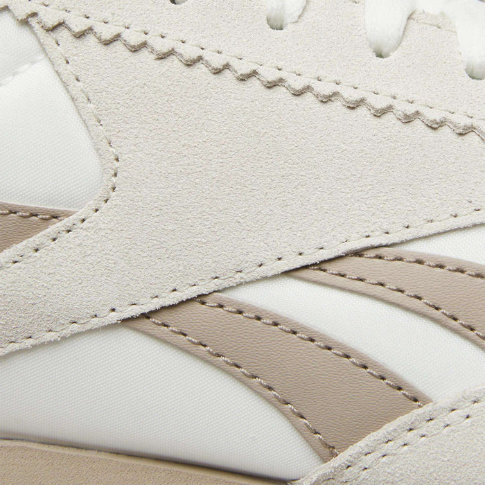 Close-up of Reebok R100 Shoes highlighting beige suede overlays, white textile uppers, and taupe synthetic stripes with detailed stitching. These low-profile sneakers feature layered textures and wavy edges inspired by '70s running styles.
