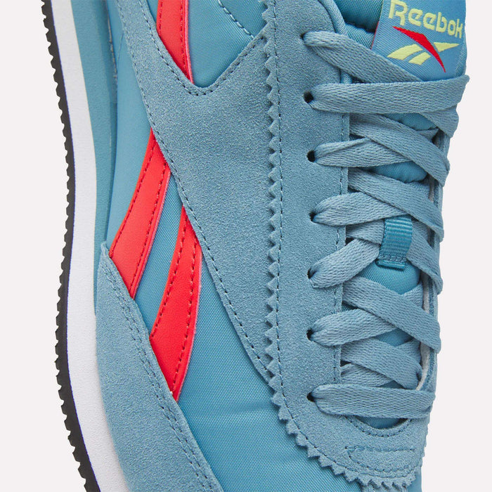 Close-up of Reebok R100 Shoes in blue with red side stripes, featuring a suede and fabric upper, blue laces, and the Reebok logo on the tongue. A black and white low-profile sole completes this '70s running-inspired sneaker.