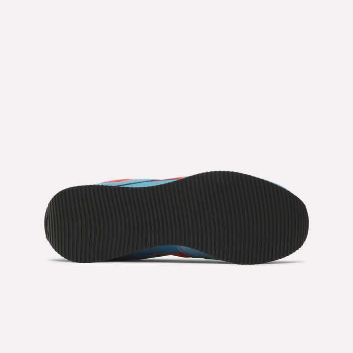 The image shows the bottom view of Reebok R100 Shoes with a black, textured rubber sole. Hints of blue and red on the upper echo '70s running style, while the plain white background highlights these low-profile sneakers.