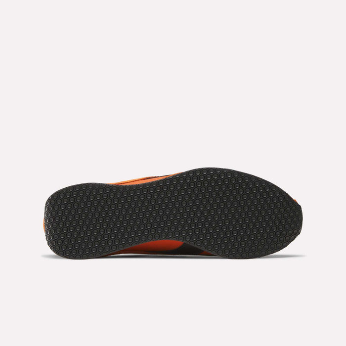 The image shows the bottom view of Reebok R400 Shoes, featuring a black rubber sole with a hexagonal pattern. The orange-edged upper and retro track-inspired, low-profile design stand out against a plain white background.