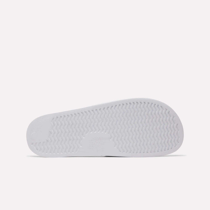 The image shows the bottom of a white Reebok RBK Fulgere Swim Slides sole with a zigzag tread for grip, a smooth circular heel, and a rectangular center section. The plain white background highlights the design.