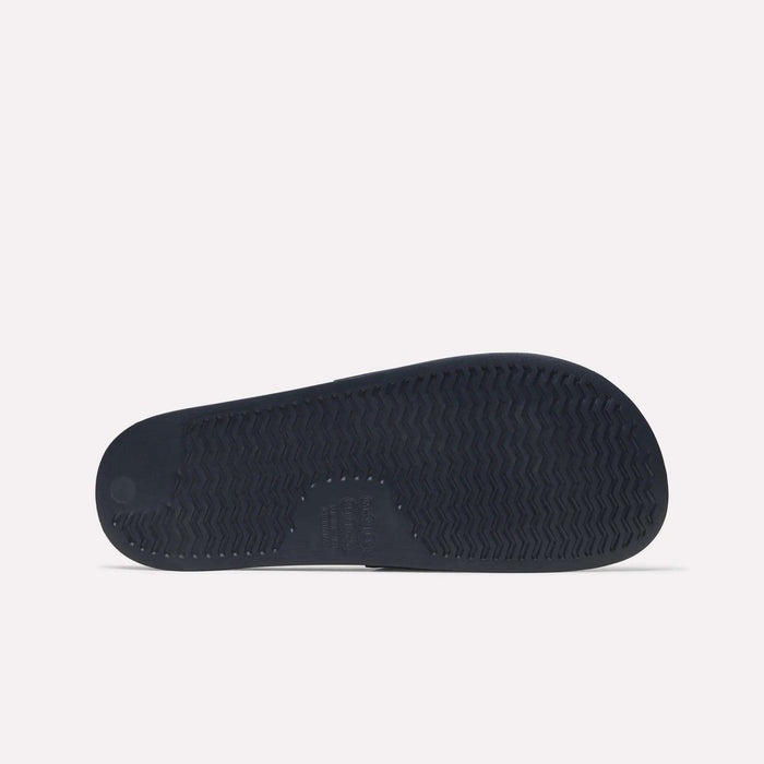 The image shows the bottom view of a dark-colored Reebok RBK Fulgere Swim Slides sole with a textured tread pattern, set against a plain white background.