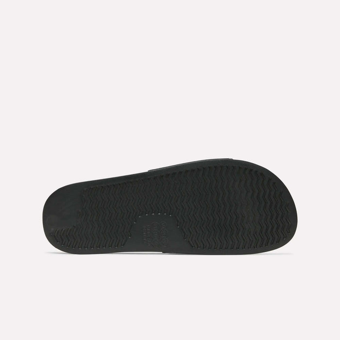 The image features the black rubber sole of the Reebok RBK Fulgere Swim Slides, shown flat with a zigzag tread and imprinted text at the arch. The slip-on sandal’s sole stands out clearly against a plain white background.