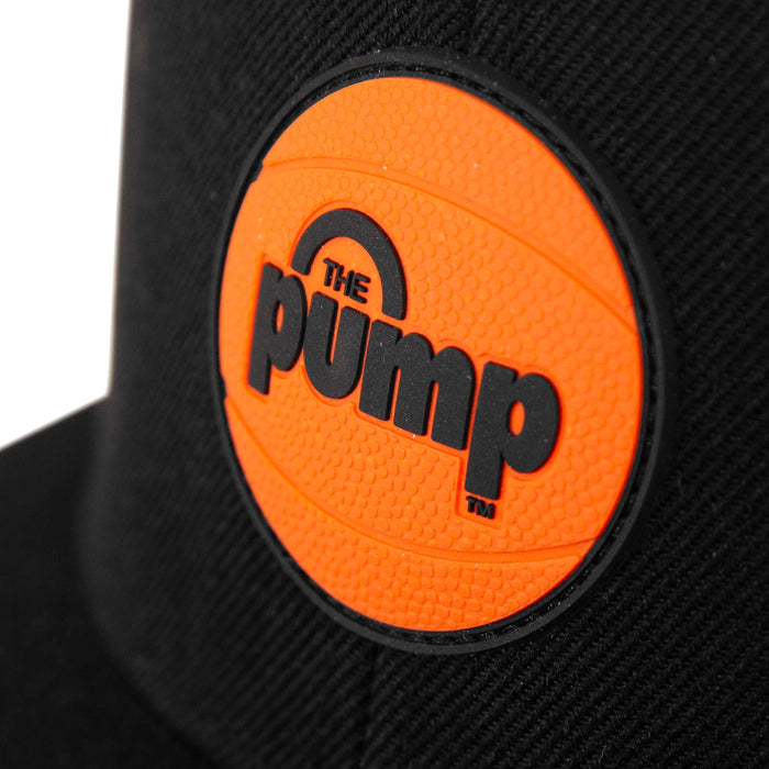 A close-up of the Reebok Pump Hat, a black cap by Reebok, featuring an orange basketball patch with "THE PUMP" in bold black letters on the front.