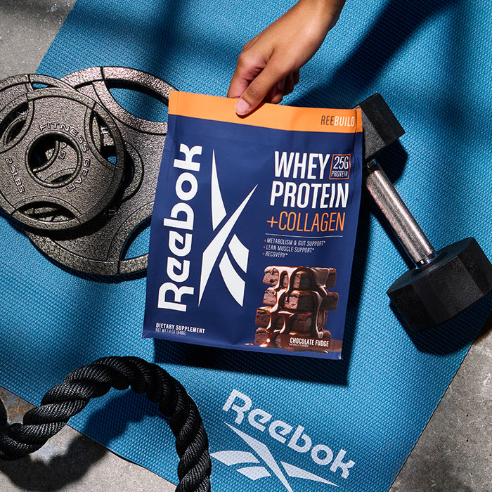 A hand holds a Reebok ReeBuild Whey Protein + Collagen chocolate fudge pouch on a blue Reebok mat, surrounded by a black dumbbell, weight plates, and rope—ideal for supporting muscle recovery.