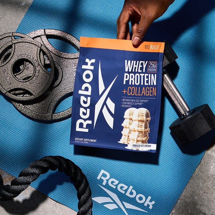 A hand holds a Reebok ReeBuild Whey Protein + Collagen packet over a blue exercise mat, surrounded by weight plates, a dumbbell, and a battle rope—ideal essentials from Reebok for muscle recovery with added collagen.