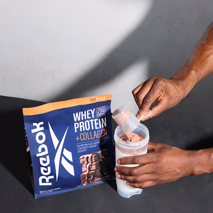 A person scoops Reebok ReeBuild Whey Protein + Collagen from a Reebok bag into a shaker bottle partly filled with liquid on a dark surface, preparing it for muscle recovery.