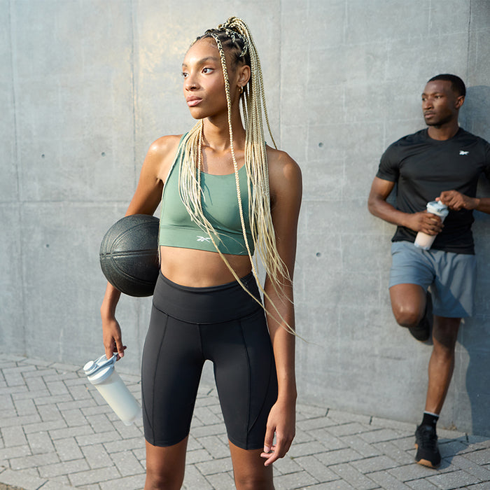 A woman in athletic wear holds a basketball and Reebok ReeBuild Whey Protein + Collagen, ready for muscle recovery, while a man in sports clothes stands behind her with a water bottle, both energized by Reebok’s performance nutrition.