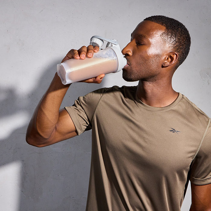 A man in a taupe athletic shirt drinks Reebok ReeBuild Whey Protein + Collagen from a plastic shaker bottle against a plain light gray background, supporting muscle recovery.